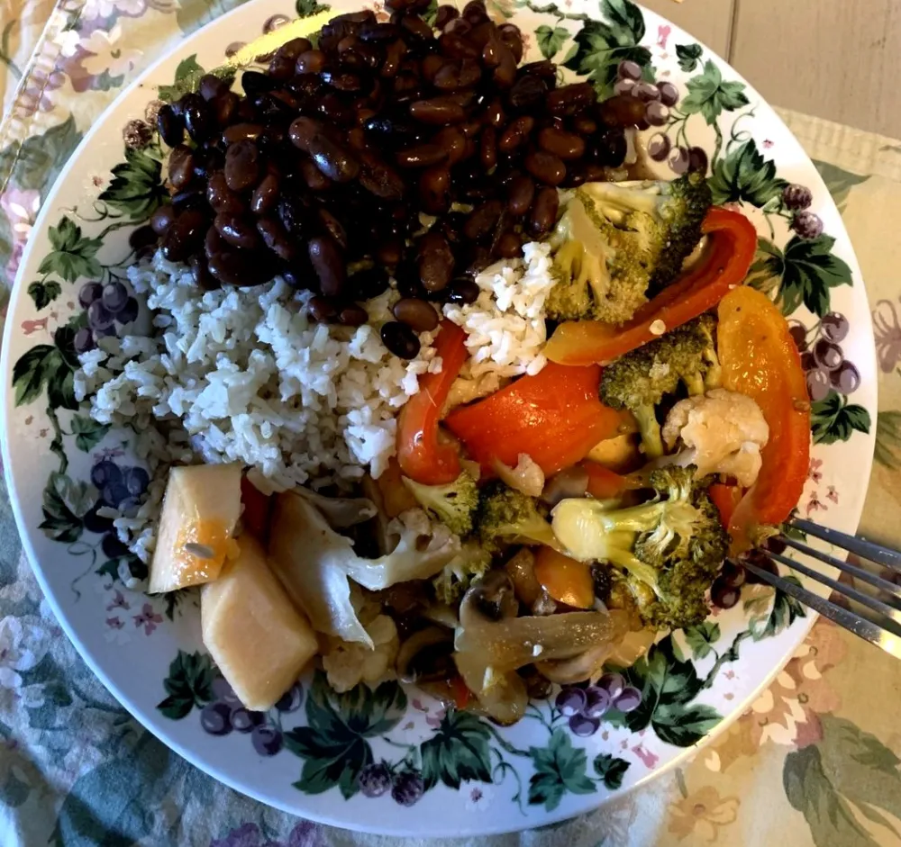 Overhead photo of a dinner plate with a floral grape-vine border, filled with brown rice, black beans, roasted broccoli, cauliflower, red peppers and mushrooms, and a few chunks of melon. A fork rests on the right edge.