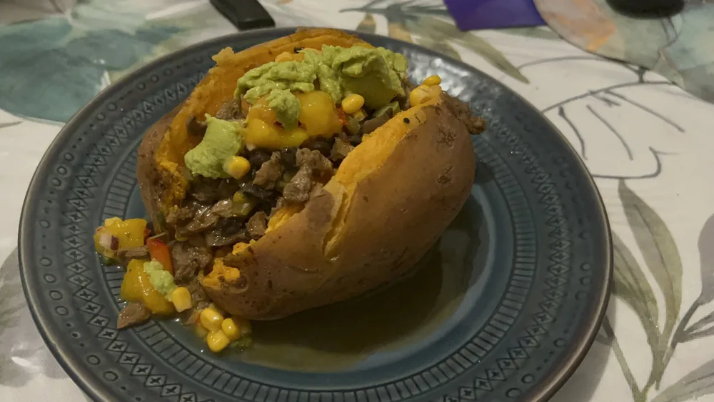 A steamed sweet potato split open on a blue patterned plate, stuffed with seasoned TVP and topped with mango salsa, corn, and mashed avocado.