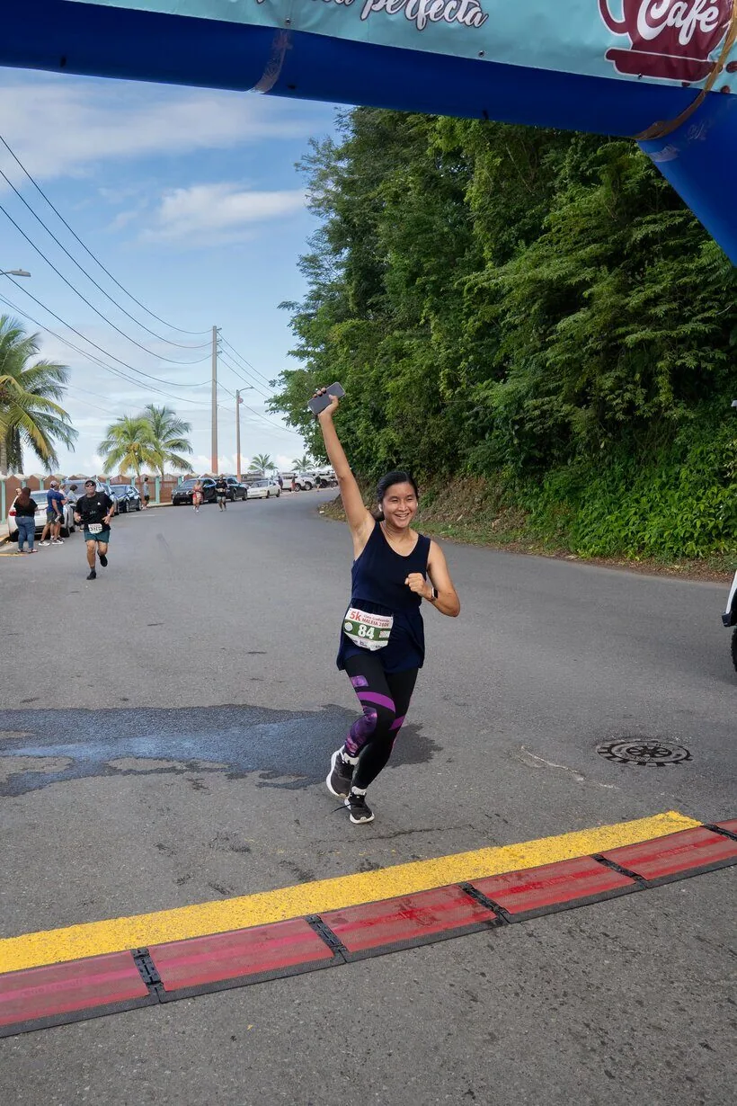 Mujer con número de corredor cruza la meta de un 5K con una mano levantada en celebración, cerca de la costa en Puerto Rico.