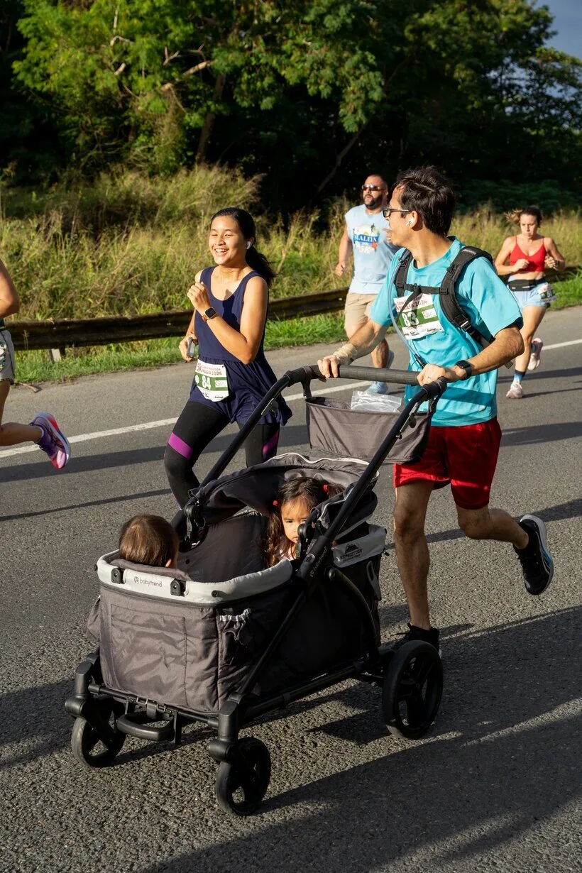 Mujer corriendo en una carrera 5K junto a otros corredores mientras un hombre empuja a dos niñ@s en un cochecito, con árboles y carretera en Puerto Rico.
